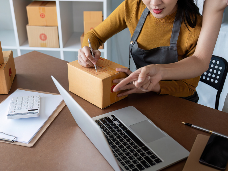 Worker labeling a shipping box while using a laptop nearby