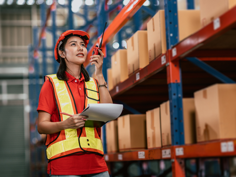 Female worker inspecting inventory with clipboard and walkie-talkie in warehouse