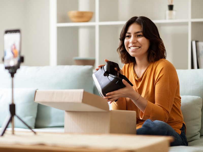 A smiling woman films herself unboxing a VR headset while sitting on a couch